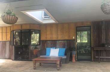 This image shows a rustic covered patio with wooden walls, a skylight, wicker sofa with blue pillows, coffee table, potted plants, hanging baskets, and glass door.