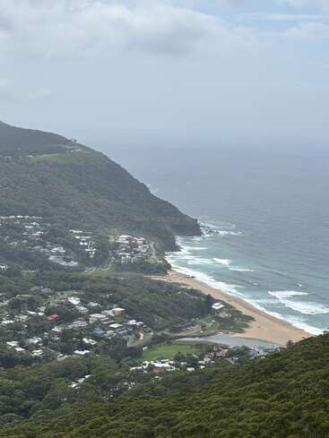 Esta imagem mostra uma cidade litorânea situada entre colinas verdes e o oceano. As casas se aglomeram perto da costa, com as ondas quebrando na praia de areia lá embaixo.