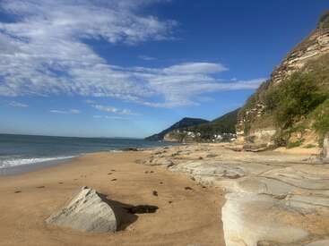 Uma bela paisagem costeira com areia dourada, falésias rochosas, ondas azuis do mar, nuvens brancas espalhadas e colinas verdes ao longe, com algumas casas visíveis. Um clima tranquilo.