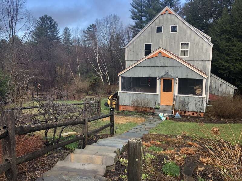 The image depicts a two-story house with a screened-in porch, surrounded by a garden and a stone pathway leading to the front door.