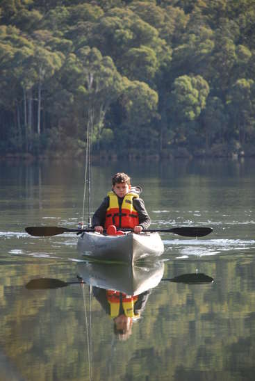 Un joven con chaleco salvavidas está sentado en un kayak en un lago, con un remo en la mano y una caña de pescar detrás, rodeado de árboles.