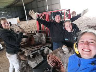 La imagen muestra a cuatro personas con protección auditiva y guantes, posando para un selfie en un almacén con madera y maquinaria de fondo.