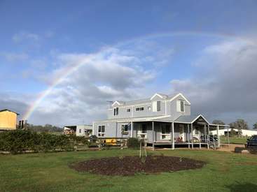 La imagen representa una casa de dos plantas con un gran porche, situada sobre un césped, con un arco iris en el cielo, sobre un fondo nublado.
