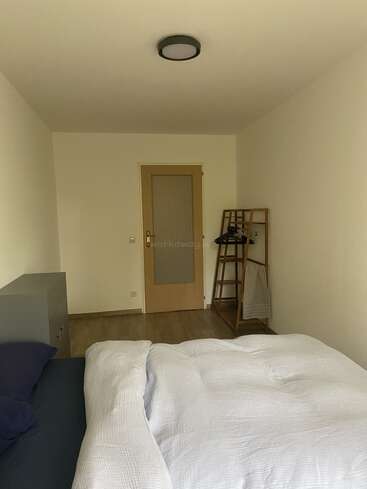 Dieses Bild zeigt ein schlichtes, aufgeräumtes Schlafzimmer mit einem gemachten Bett, einer Holztür, einer Deckenlampe, einem Schrank und einem Kleiderständer aus Holz.