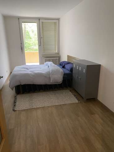 Ein schlichtes Schlafzimmer mit Holzboden, Einzelbett, weißer Bettwäsche, blauem Kissen, Metallschrank, kleinem Teppich, großem Fenster, Balkon und Tageslicht.