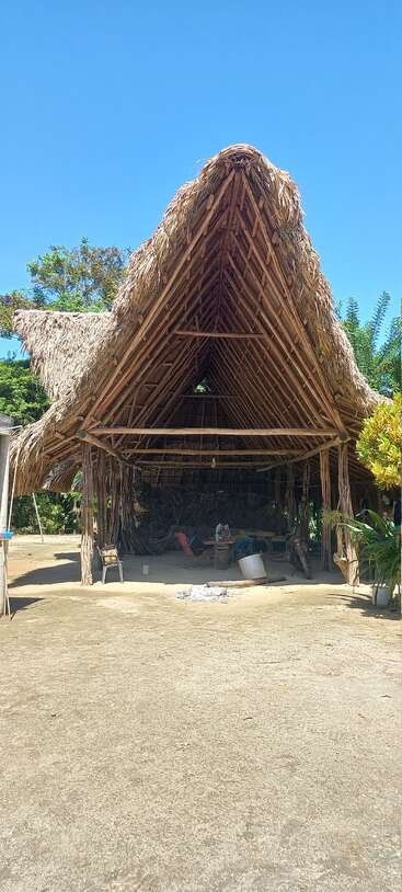 Eine große strohgedeckte Hütte aus Holz und Palmblättern steht unter einem klaren blauen Himmel, in der Stühle, ein Motorrad und Werkzeuge zu sehen sind.