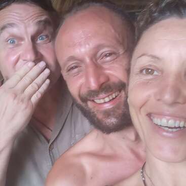 Trois adultes sourient de près pour prendre un selfie. L'homme de gauche se couvre la bouche d'un air amusé, tandis que les autres sourient joyeusement, rayonnant de bonheur et d'amitié.