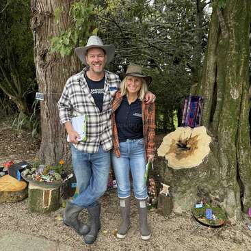 A smiling man and woman pose outdoors by large trees, wearing hats, plaid shirts, and boots. They hold notebooks, surrounded by whimsical miniature fairy gardens.