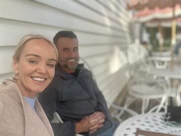 Eine lächelnde Frau und ein lächelnder Mann sitzen zusammen draußen in einem Café mit weißen Stühlen und Tischen und genießen einen schönen Tag. Im Hintergrund ist eine weiße Holzwand zu sehen.