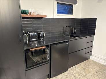 This modern kitchenette features black cabinets, sleek countertops, a small fridge, microwave, toaster, kettle, open shelving with glassware, black tiled backsplash, and a stainless sink.