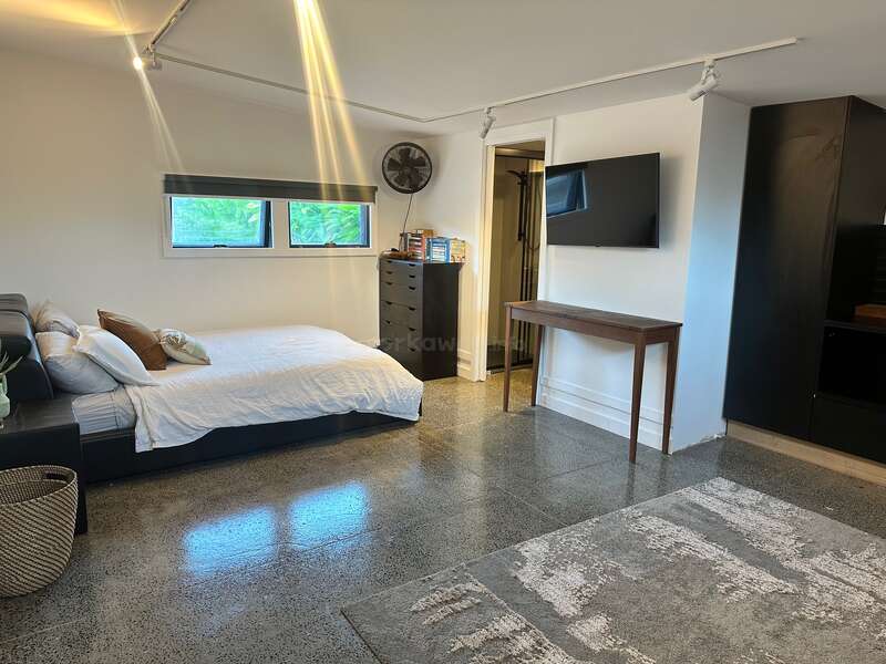 A imagem mostra um quarto moderno com cama, cômoda, TV e escrivaninha, com paredes brancas, piso de concreto polido e uma grande janela com vista para a vegetação externa.