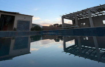 Uma piscina serena reflete o céu azul, as nuvens e dois edifícios rústicos ao anoitecer. A água calma cria uma imagem espelhada perfeita, exalando tranquilidade.