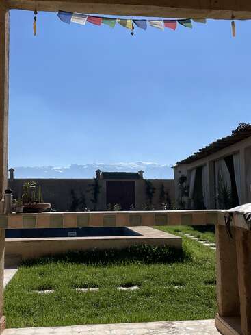 Ein ruhiger Innenhof mit grünem Gras, einem Pool in der Mitte und bunten Gebetsfahnen darüber. Unter dem klaren blauen Himmel sieht man Berge.