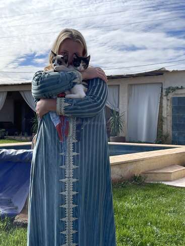 Eine Frau in einem blauen traditionellen Kleid steht draußen an einem Pool und knuddelt zwei süße Kätzchen, mit einem gemütlichen Haus und blauem Himmel im Hintergrund.