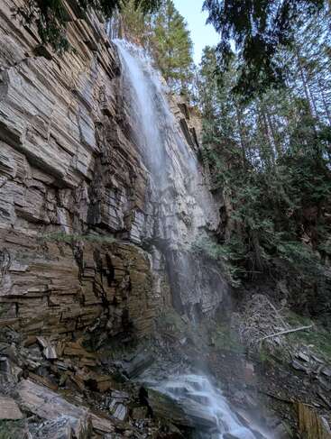 A stunning waterfall cascades down layered rocky cliffs, surrounded by dense green trees. Mist rises at the base, creating a serene, natural scene in the forest.