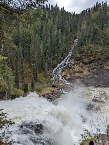 Uma poderosa cachoeira cai em cascata sobre as rochas em uma piscina cheia de névoa, cercada por uma densa floresta sempre verde e terreno acidentado, com céu nublado, criando uma cena serena na natureza.
