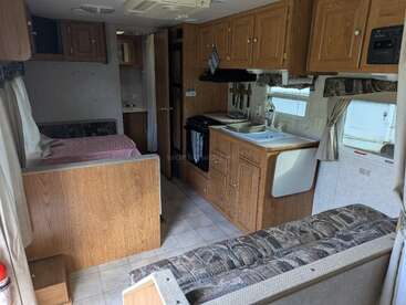 Esta imagem mostra o interior de um trailer, com uma pequena cozinha, área de estar, mesa, cama na parte de trás, armários de madeira e piso de azulejos. Espaço compacto e aconchegante.