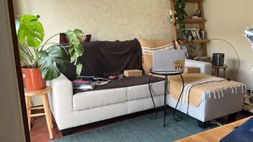 A imagem mostra uma sala de estar com um sofá, uma mesa de centro e uma TV, com uma grande janela e uma lareira, com uma estante e uma parede decorativa ao fundo.