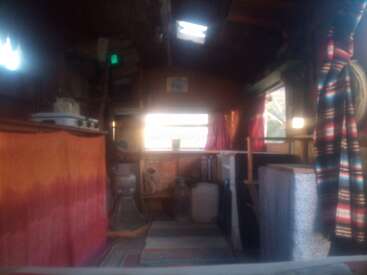 Um quarto aconchegante e rústico com cortinas e tapete coloridos, pequena área de cozinha, prateleiras e luz do sol entrando pela janela. Ambiente acolhedor, convidativo, simples e caseiro.