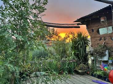 A rustic clay house sits amid a lush, green garden filled with potted plants. The vibrant sunset sky glows orange and yellow, creating a peaceful atmosphere.