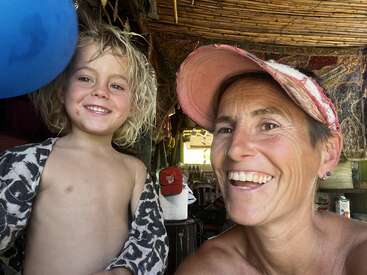 A happy woman wearing a pink cap smiles alongside a young, shirtless, blonde-haired child, both in a rustic, colorful, and cozy indoor setting.