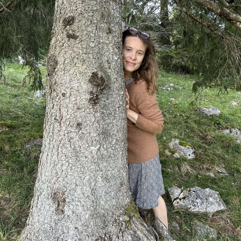 Une femme se tient derrière un gros tronc d'arbre dans une forêt verdoyante. Elle sourit doucement, vêtue d'un pull marron, d'une jupe à motifs et de lunettes de soleil posées sur la tête.