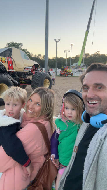 A smiling family of four poses at a monster truck event. Two young children are held by parents. Monster trucks and event equipment are visible in the background.