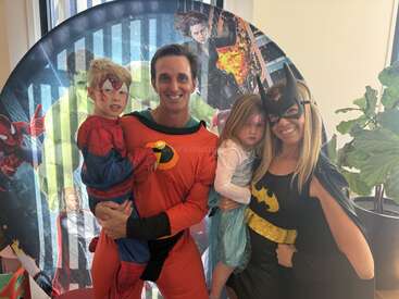 A family dressed as superheroes poses happily. The boy is Spider-Man, the girl is Elsa, dad is Mr. Incredible, and mom is Batgirl. Colorful, joyful background.