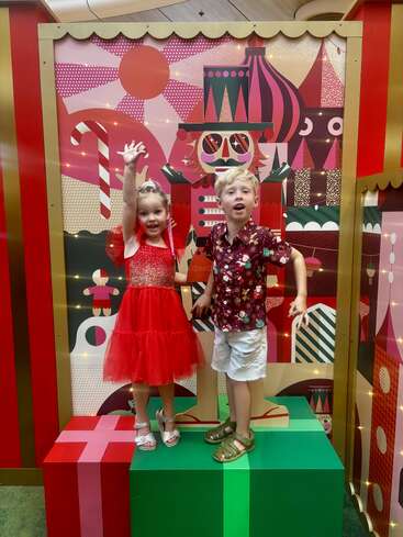 Two children happily pose on large colorful gift boxes, wearing festive clothing, in front of a whimsical, vibrant Christmas-themed backdrop featuring a nutcracker and candy canes.