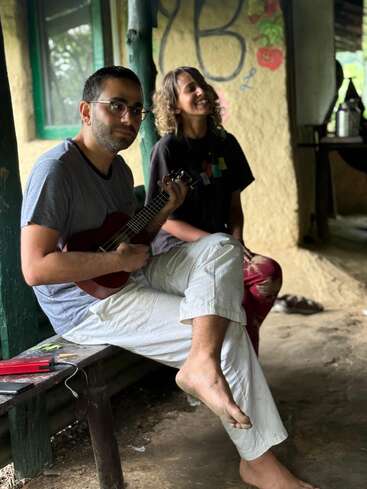 Un homme est assis pieds nus sur un banc et joue du ukulélé, tandis qu'une femme souriante profite de l'instant à ses côtés, dans un cadre rustique et artistique.