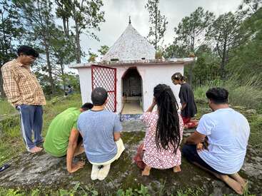 Un groupe de personnes s'agenouille et prie devant un petit temple blanc entouré de verdure et d'arbres, créant une atmosphère extérieure paisible et spirituelle.
