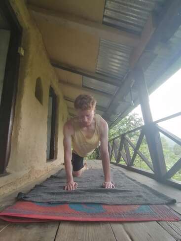 Une personne aux cheveux bouclés pratique le yoga ou l'exercice sur un tapis en plein air, sous un porche rustique en bois, entouré de nature et de verdure, sous un toit en tôle.