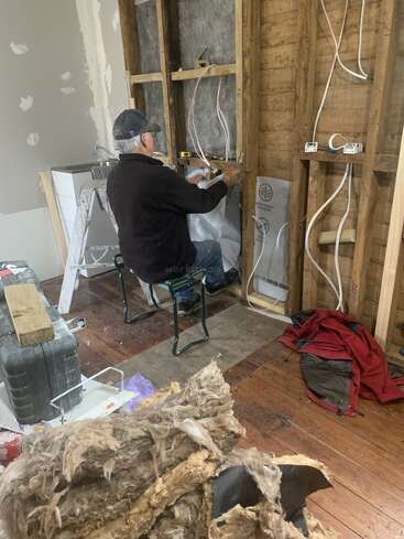 A man works on exposed wall wiring during a home renovation. Insulation lies on the floor, tools and materials are scattered, and unfinished surfaces surround him.