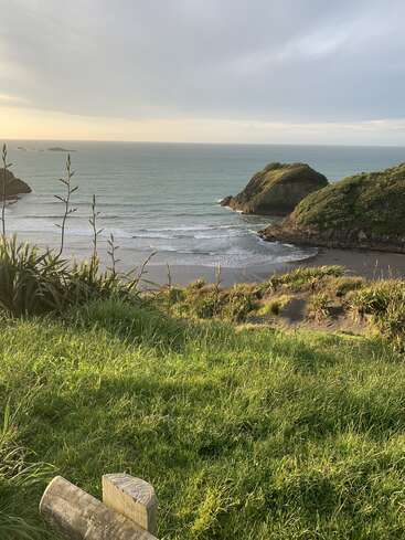 A scenic coastal view with lush green grass, sandy beach, and rocky outcrops. Gentle ocean waves roll in under a cloudy sky at sunset.