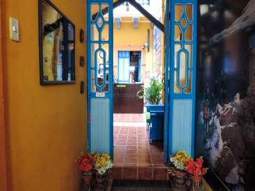 Un couloir étroit aux murs orange et bleus mène à une cour intérieure. Des portes bleues, des pots de fleurs, des œuvres d'art et une lumière naturelle éclatante créent une atmosphère accueillante.