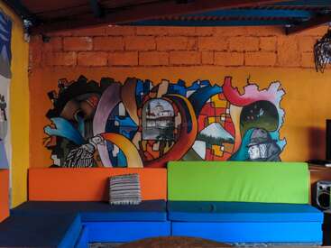 Un mur salon dynamique présente des peintures murales abstraites et culturelles colorées au-dessus d'un canapé moderne multicolore avec un seul coussin rayé, créant une atmosphère artistique et chaleureuse.