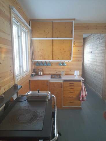 The image depicts a compact kitchen with light-colored wood paneling, featuring a stove, cabinets, and a window, with a doorway leading to another room.