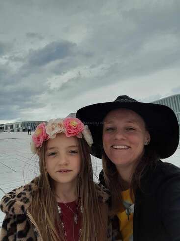 The image depicts a woman and a young girl posing for a photo, with the woman wearing a black hat and the girl adorned with a pink flower crown.