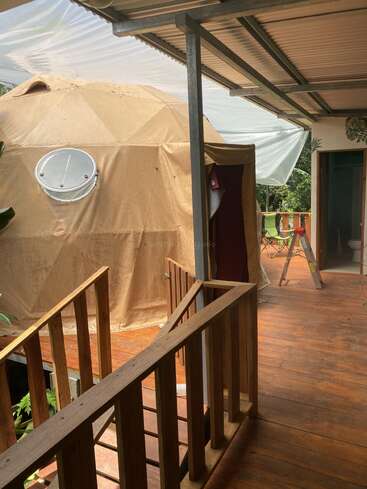 Sobre una terraza de madera, parcialmente cubierta por un tejado de chapa ondulada y rodeada de vegetación tropical, se levanta una tienda de campaña en forma de cúpula geodésica con una ventana circular.