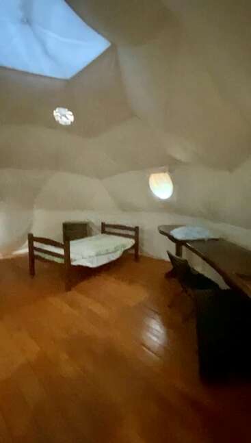 Esta imagen muestra una acogedora habitación minimalista con una cama individual, suelo de madera, un pequeño escritorio, una silla y ventanas redondas en forma de ojo de buey en una estructura de cúpula.
