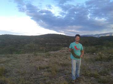 A man stands in a field, holding a wreath, wearing a turquoise t-shirt with a red bird design, in front of a mountainous landscape under a cloudy sky.