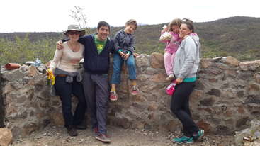 The image shows a group of people posing for a photo, likely a family, with a mix of adults and children, all dressed in casual clothing, posing together in a relaxed outdoor setting.