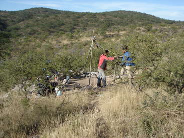 Two men work together to pull up a rope from a well in a desert landscape, surrounded by bushes and dry grass, with a hill in the background.