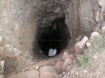 The image depicts a cave with a dark interior, featuring a hole in the center and rocks surrounding it, set against a natural stone background.