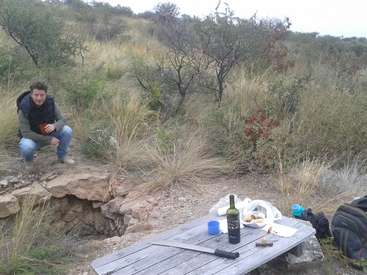 The image depicts a man squatting on a rocky outcrop, with a picnic table featuring wine, food, and a knife in the foreground, set against a natural outdoor backdrop.