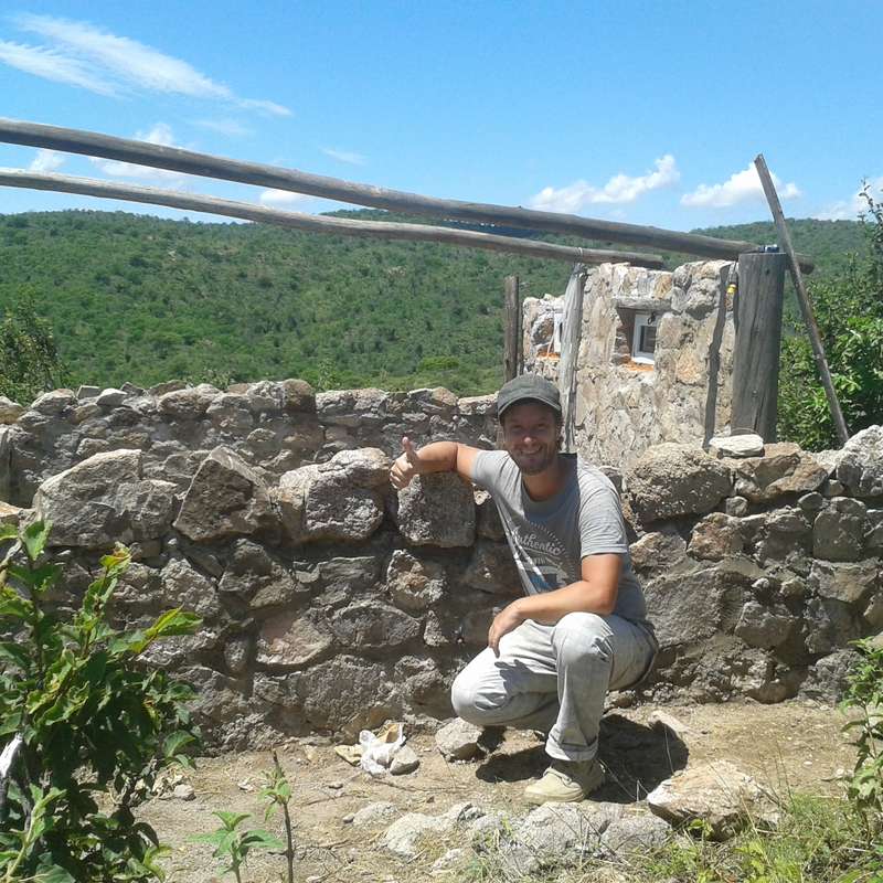 The image depicts a man posing in front of a stone wall, with a rustic wooden structure and a lush green hillside under a clear blue sky with scattered clouds.