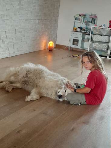 Une jeune fille aux cheveux longs est assise par terre et caresse un gros chien blanc dans une pièce où se trouvent une cuisine et des jouets éparpillés.