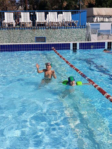 L'image représente deux enfants dans une piscine, l'un portant un bonnet de bain vert et l'autre un flotteur vert, tous deux dans l'eau.