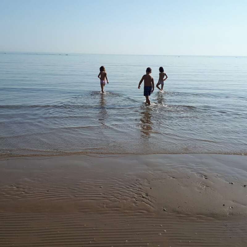 Trois enfants pataugent dans l'océan depuis une plage de sable, leurs silhouettes se détachant sur l'eau calme et le ciel clair d'une journée ensoleillée.