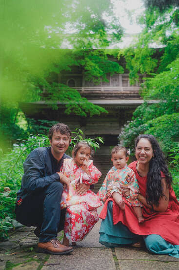 A smiling family of four poses outdoors surrounded by lush greenery. The children wear colorful traditional attire, and a beautiful wooden building is visible in the background.